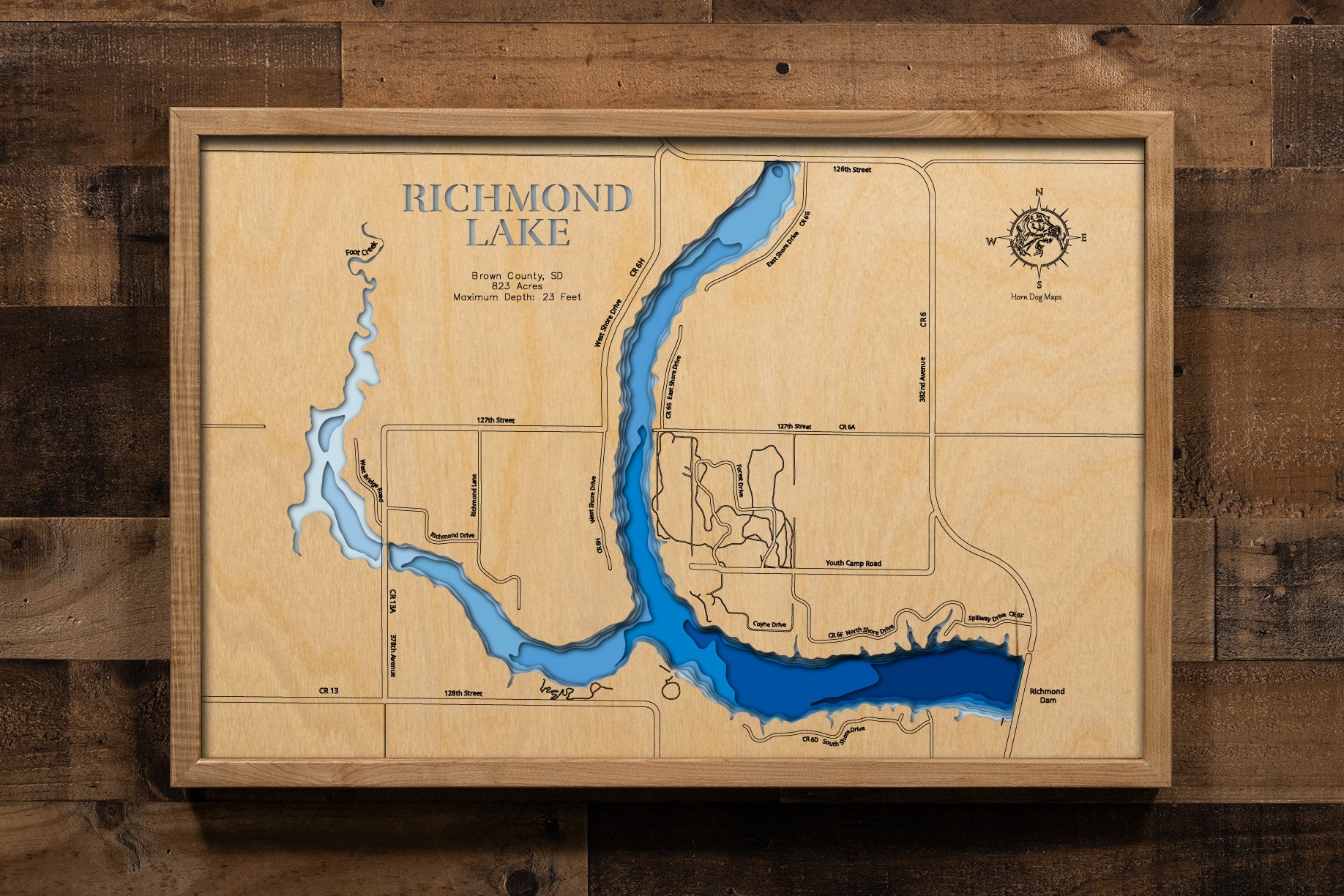 A finely crafted, laser-cut, multi-layered wooden topographic map of Richmond Lake in Brown County, South Dakota. The dimensional relief showcases the 823-acre WPA-era reservoir's bathymetric features from the earthen dam to the shallow western arm where Foote Creek enters, depth contours revealing the 23-foot maximum depth near the dam, and the three public access points.