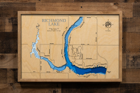 A finely crafted, laser-cut, multi-layered wooden topographic map of Richmond Lake in Brown County, South Dakota. The dimensional relief showcases the 823-acre WPA-era reservoir's bathymetric features from the earthen dam to the shallow western arm where Foote Creek enters, depth contours revealing the 23-foot maximum depth near the dam, and the three public access points.
