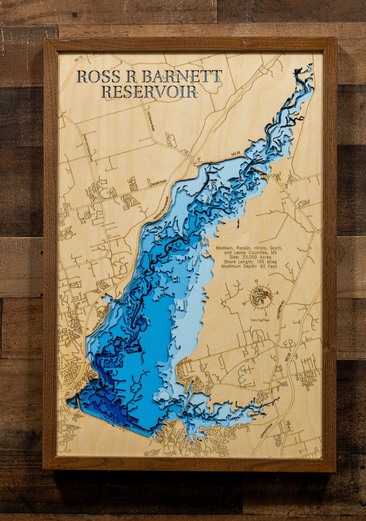 A framed wooden depth map of Ross R Barnett Reservoir, featuring layered blue and light blue water levels on a light wood background, mounted on a dark wood plank wall.