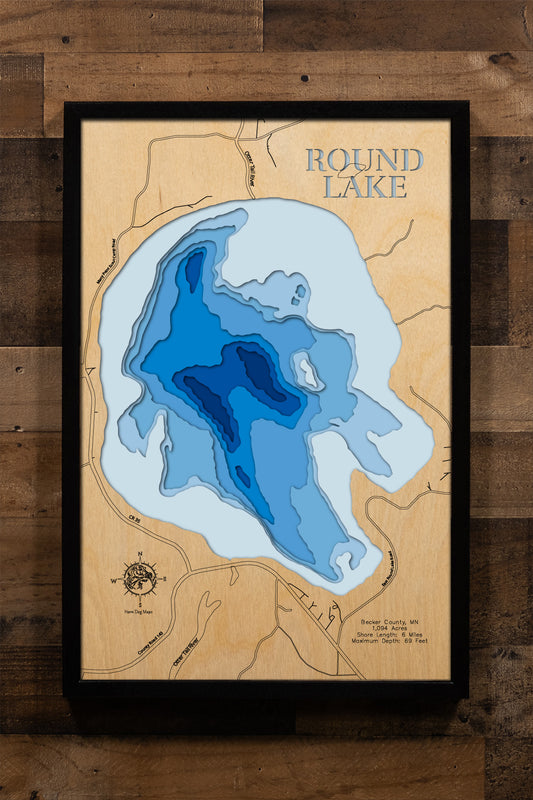 A precision laser-cut, multi-layered wooden map of Round Lake in Becker County, Minnesota, within the White Earth Indian Reservation near the Tamarac National Wildlife Refuge. The natural wood top layer captures 6 miles of shoreline in fine detail, while a light-to-dark blue gradient highlights the lake's notable 69-foot depth. A handcrafted statement piece for any Minnesota lake cabin, Northwoods home, or outdoor enthusiast's wall.