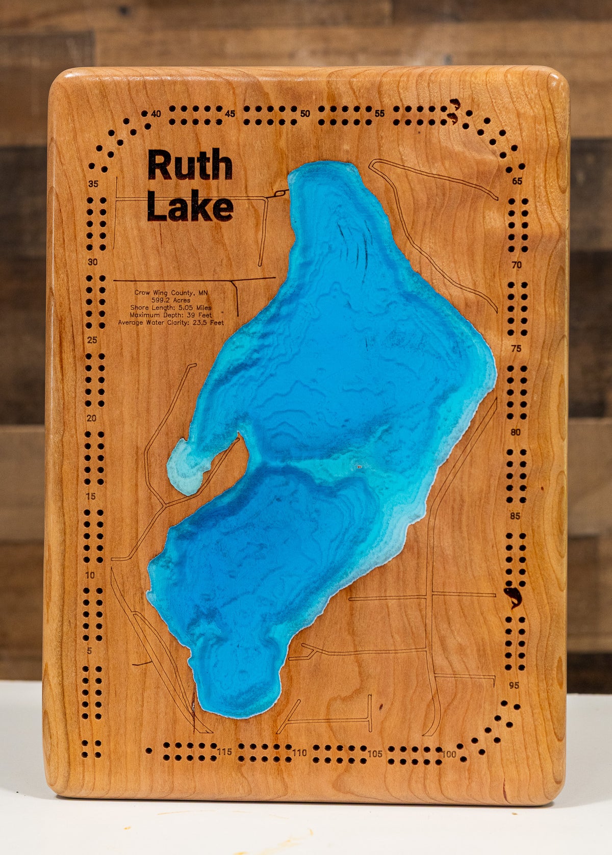 A cribbage board featuring a 3D resin map of 'Ruth Lake' in bright blue, set against a warm honey-toned wood grain background with pre-drilled holes around the border.