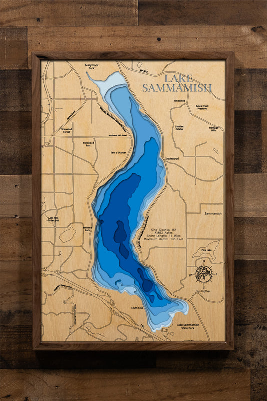 A magnificently detailed, laser-cut, multi-layered wooden topographical map of Lake Sammamish in King County, Washington, showcasing the pristine 4,853-acre lake stretching seven miles from Issaquah to Redmond with dramatic depth contours from the 29-foot average to the 105-foot maximum depth. The natural wood top layer frames 11 miles of shoreline including Marymoor Park, Lake Sammamish State Park with nine boat launches, the East Lake Sammamish Trail, Issaquah Creek inlet, and Sammamish River outlet, whil