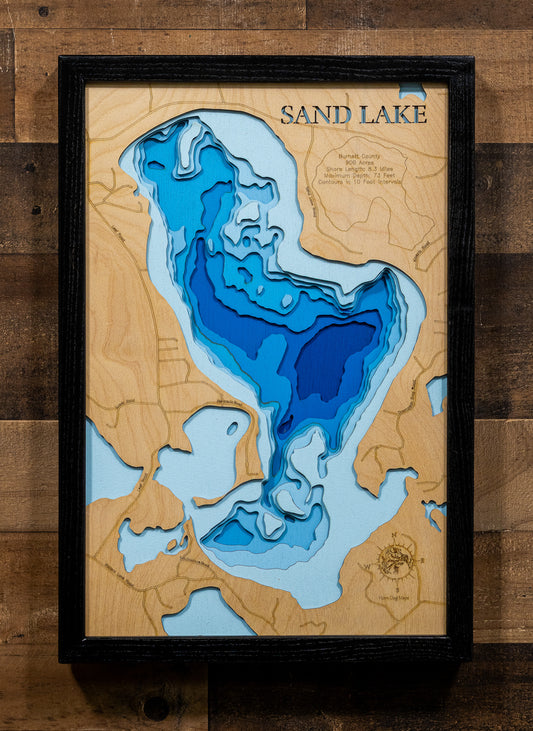 A framed 3D wooden depth map of Sand Lake, featuring layered wood sections in shades of blue, light blue, and natural wood grain, mounted on a rustic dark wood wall.