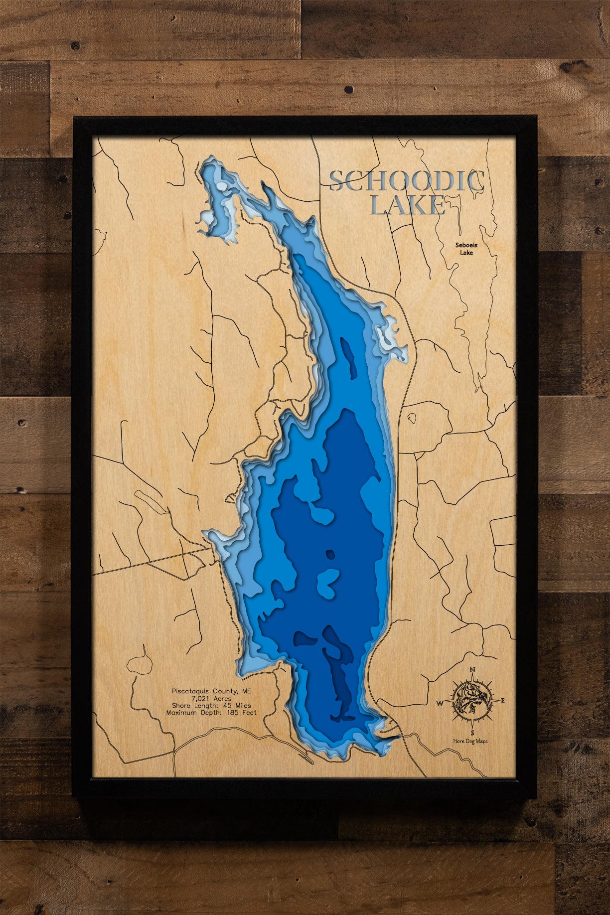 A magnificently detailed, laser-cut, multi-layered wooden topographical map of Schoodic Lake in Piscataquis County, Maine, showcasing the expansive 7,021-acre deep glacial lake with dramatic depth contours from shallow tributary areas to the astounding 185-foot maximum depth. The natural wood top layer frames approximately 45 miles of shoreline covering Lake View Plantation and Brownville, Schoodic Stream outlet, and two public boat launches, while graduated blue layers create a stunning visual gradient.