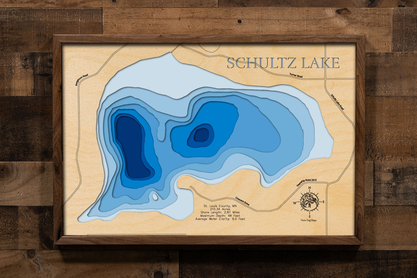 A laser-cut, layered wooden map of Schultz Lake in St. Louis County, Minnesota. The shape of the lake is visible within the northeastern Minnesota landscape. The varying depths of the lake, reaching a maximum of 48 feet, are represented by different layers of wood.