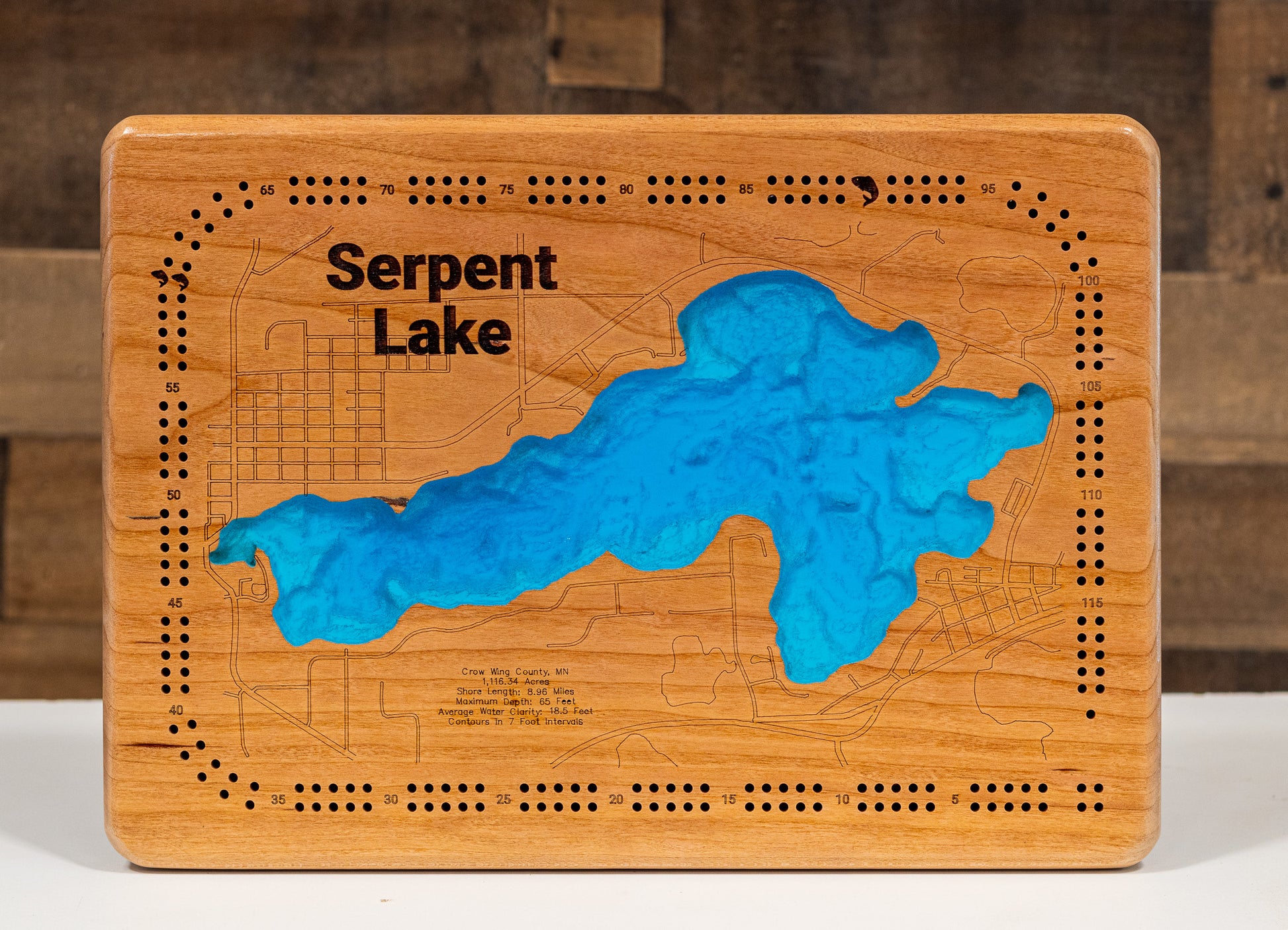 A laser-cut, layered wooden map of Serpent Lake in Crow Wing County, Minnesota. The distinct shape of this natural lake, with its 8.93-mile shoreline, is clearly visible within the forested landscape of central Minnesota near the towns of Crosby and Deerwood. The varying depths of the lake, reaching a maximum of 65 feet, are represented by different layers of wood, illustrating its impressive underwater topography.