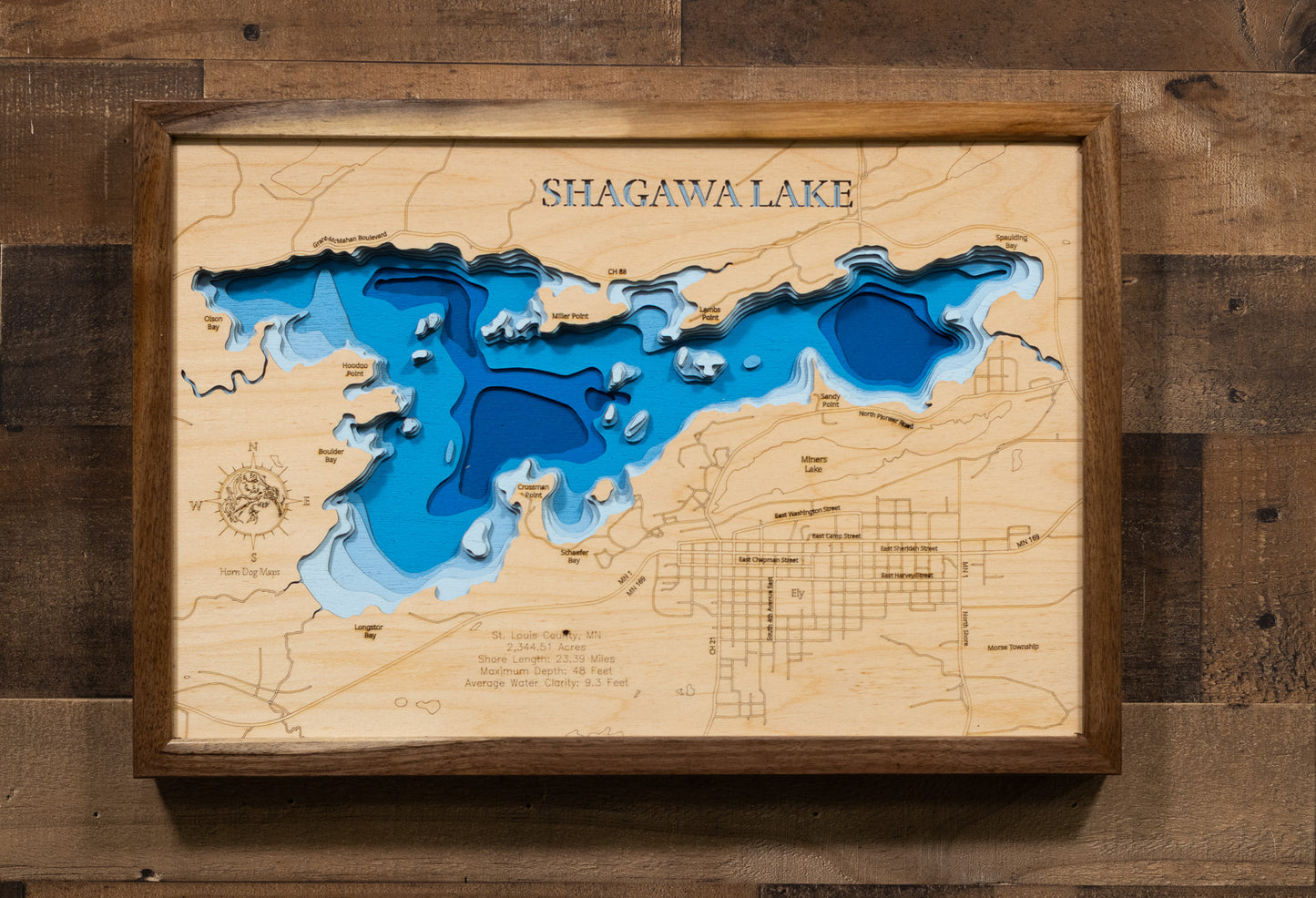 A laser-cut, layered wooden map of Shagawa Lake in St. Louis County, Minnesota. The distinct shape of this natural lake, with its 23.39-mile shoreline, is clearly visible within the forested landscape of northeastern Minnesota near the city of Ely. The varying depths of the lake, reaching a maximum of 48 feet, are represented by different layers of wood, illustrating its impressive underwater topography.