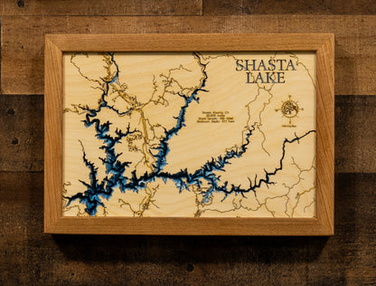 A framed wooden map of Shasta Lake, featuring a light natural wood background with dark blue water channels and engraved details, housed in a matching light wood frame and displayed against a rustic dark wood wall.