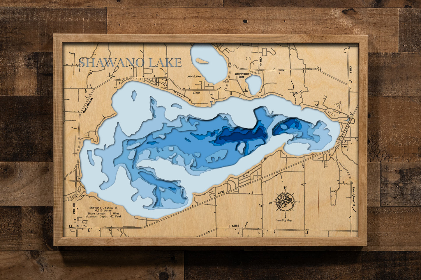A beautifully crafted, laser-cut, multi-layered wooden topographical map of Shawano Lake in Shawano County, Wisconsin, showcasing the lake's 6,215-acre surface area with detailed depth contours from the shallow 9-foot average depth to the 42-foot maximum depth. The natural wood top layer frames 18 miles of precisely rendered shoreline, while graduated blue layers create a stunning visual depth gradient.