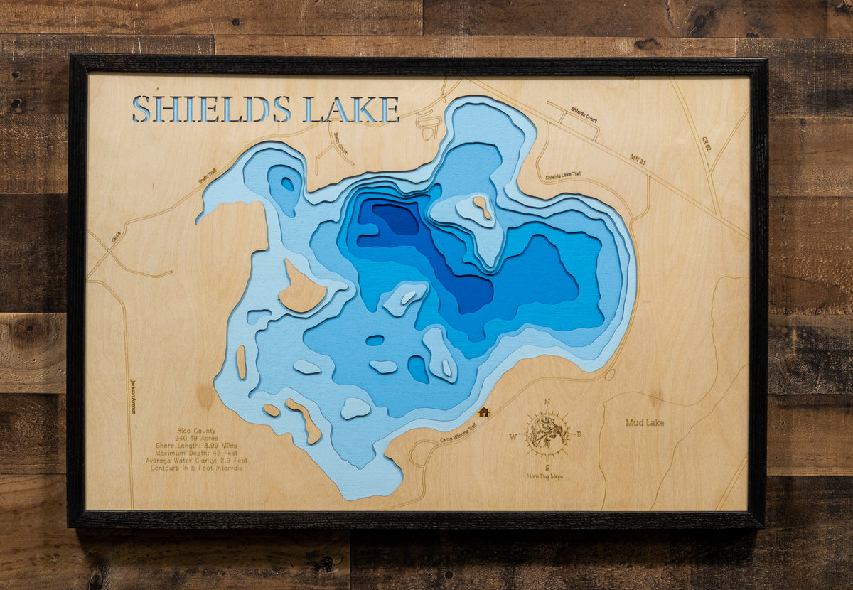 A framed wooden depth map of Shields Lake, featuring layered light and dark blue contours on a natural wood background, mounted on a dark wood plank wall.