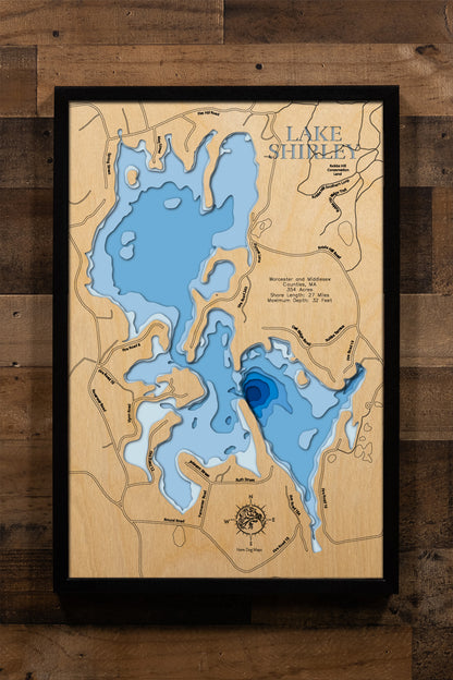 A meticulously detailed, laser-cut, multi-layered wooden topographical map of Lake Shirley in Worcester and Middlesex Counties, Massachusetts, showcasing the irregularly shaped 354-acre reservoir with an impressive 27 miles of shoreline featuring numerous coves and islands. The natural wood top layer frames the historic dam on Catacunemaug Brook, Shady Point Beach & Campground, the public boat launch, and heavily developed shoreline, while graduated blue layers create a gentle visual depth gradient from the