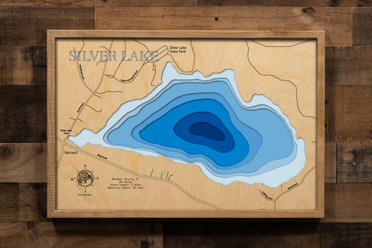 A beautifully detailed, laser-cut, multi-layered wooden topographical map of Silver Lake in Windsor County, Vermont, showcasing the intimate 84-acre warm-water lake with depth contours from shallower areas to the 32-foot maximum depth. The natural wood top layer frames approximately two miles of shoreline including Silver Lake State Park, the historic Barnard General Store village beach, and nearly 50 percent protected conservation land, while graduated blue layers create a gentle visual depth gradient.