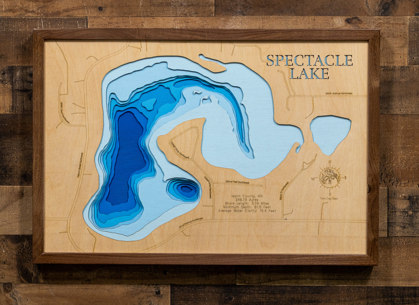 A framed wooden depth map art piece of Spectacle Lake, featuring layered blue and light blue water contours on a light wood background, enclosed in a dark wood frame against a rustic wood plank wall.