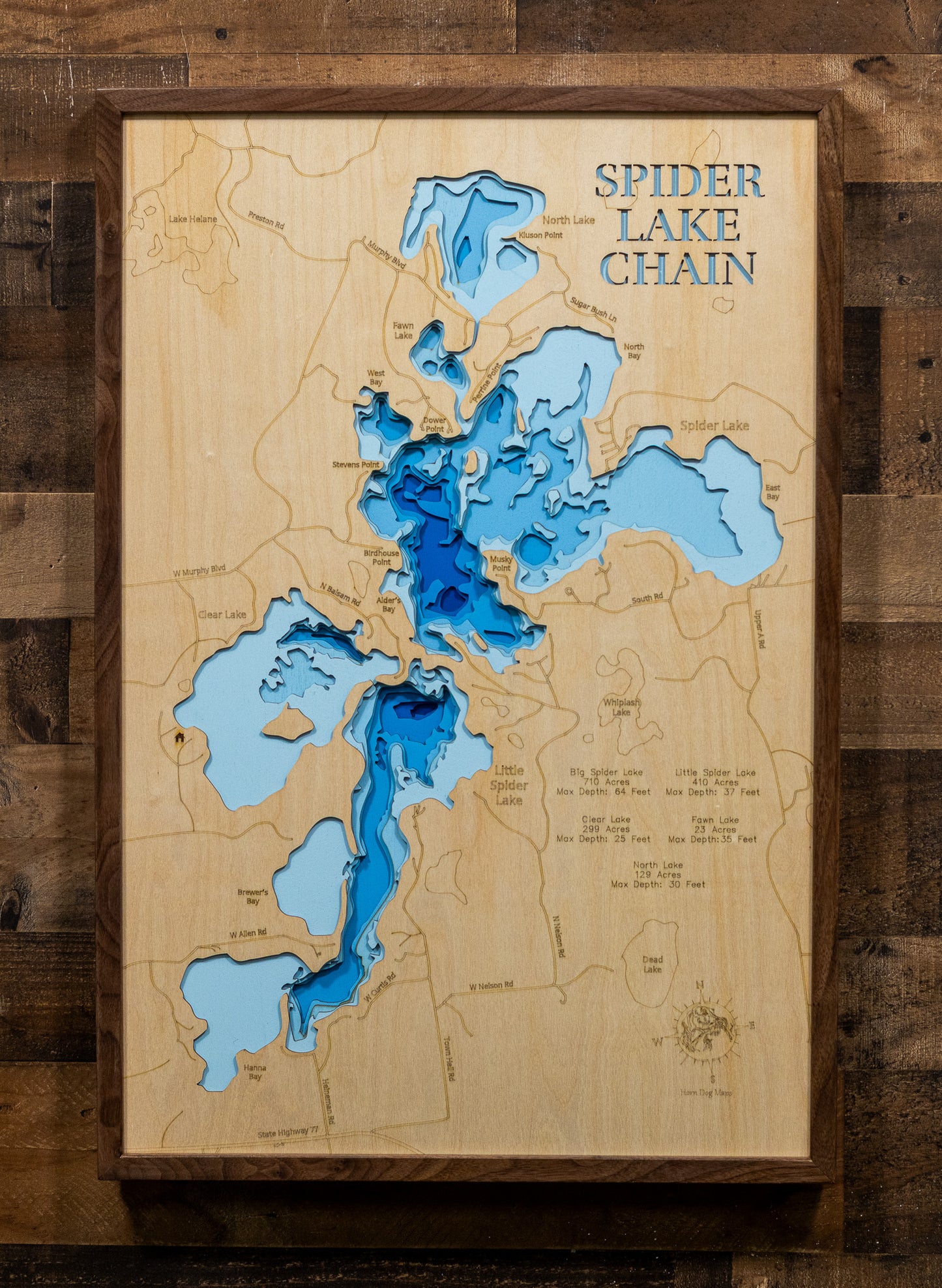 A framed 3D wooden map of the Spider Lake Chain, featuring layered blue wood cutouts representing different depths of water on a natural light wood background, mounted on a rustic dark wood plank wall.