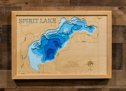 A framed wooden depth map of Spirit Lake, featuring layered blue and light blue water contours on a light wood grain background, mounted on a dark wood plank wall.