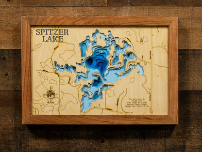 A framed wooden Spitzer Lake depth map art piece, featuring layered blue and light blue wood cutouts representing the lake's contours, mounted on a rustic dark wood background.