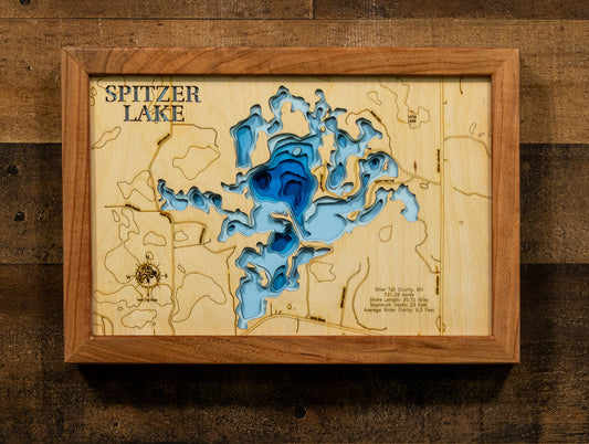 A framed wooden Spitzer Lake depth map art piece, featuring layered blue and light blue wood cutouts representing the lake's contours, mounted on a rustic dark wood background.