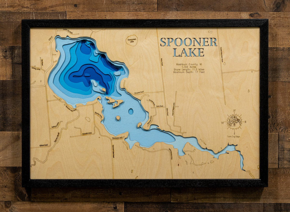 A framed wooden Spooner Lake depth map wall art, featuring layered wood cutouts in shades of blue to represent water depth, mounted on a rustic brown wood plank wall.