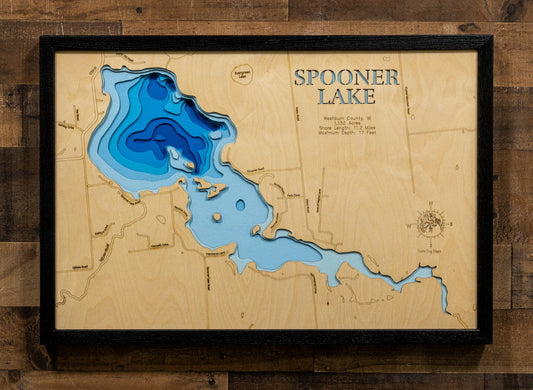 A framed wooden Spooner Lake depth map wall art, featuring layered wood cutouts in shades of blue to represent water depth, mounted on a rustic brown wood plank wall.