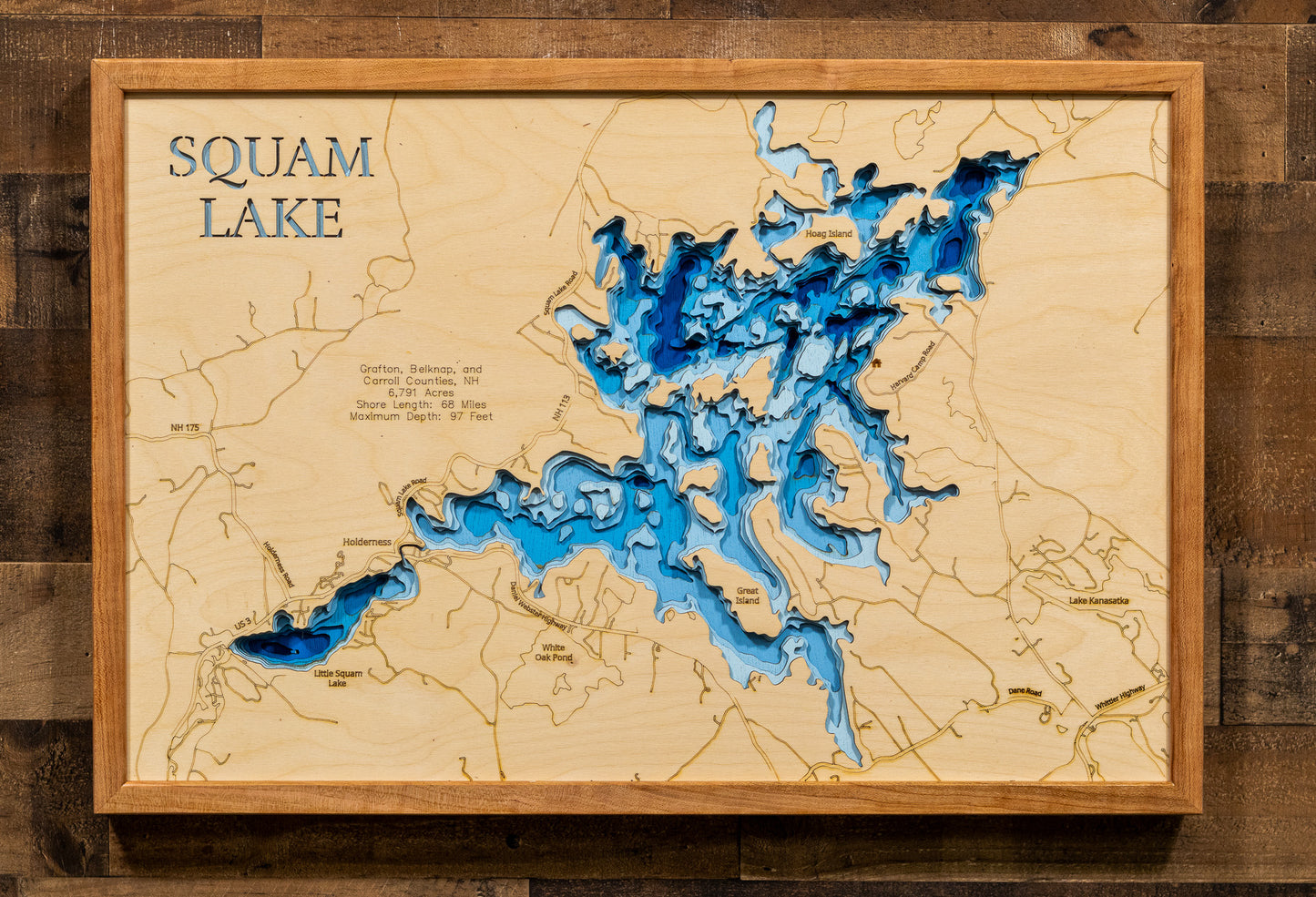 A laser-cut, layered wooden map of Squam Lake, located in Grafton, Belknap, and Carroll Counties, New Hampshire. The intricate shape of the lake, with its many islands contributing to its 68-mile shoreline, is clearly depicted within the central New Hampshire landscape. The varying depths of the lake, reaching a maximum of 97 feet, are represented by different layers of wood, creating a three-dimensional effect of this beautiful and well-known water body.