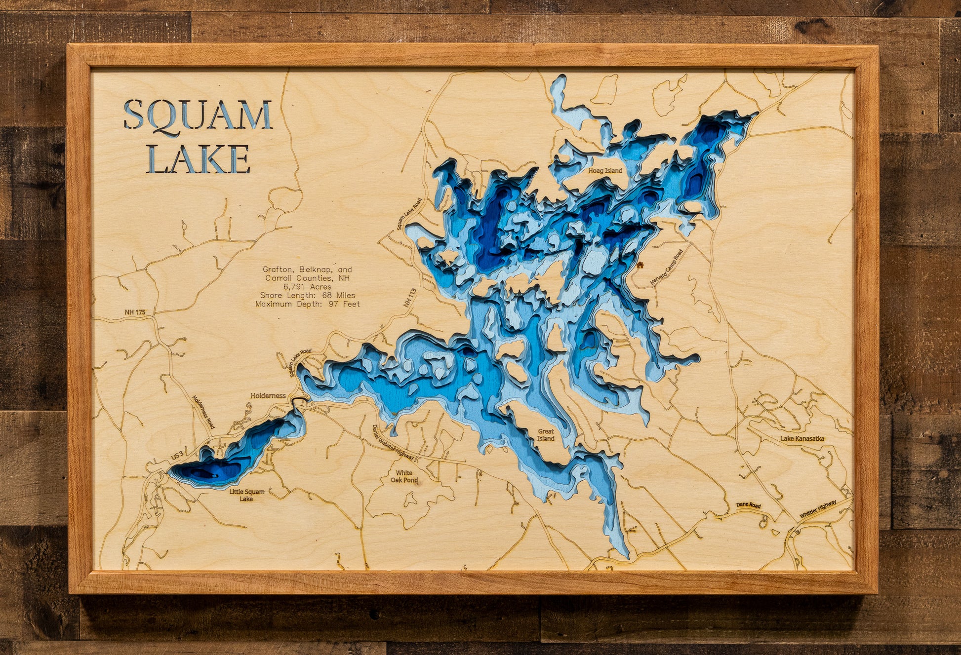 A laser-cut, layered wooden map of Squam Lake, located in Grafton, Belknap, and Carroll Counties, New Hampshire. The intricate shape of the lake, with its many islands contributing to its 68-mile shoreline, is clearly depicted within the central New Hampshire landscape. The varying depths of the lake, reaching a maximum of 97 feet, are represented by different layers of wood, creating a three-dimensional effect of this beautiful and well-known water body.