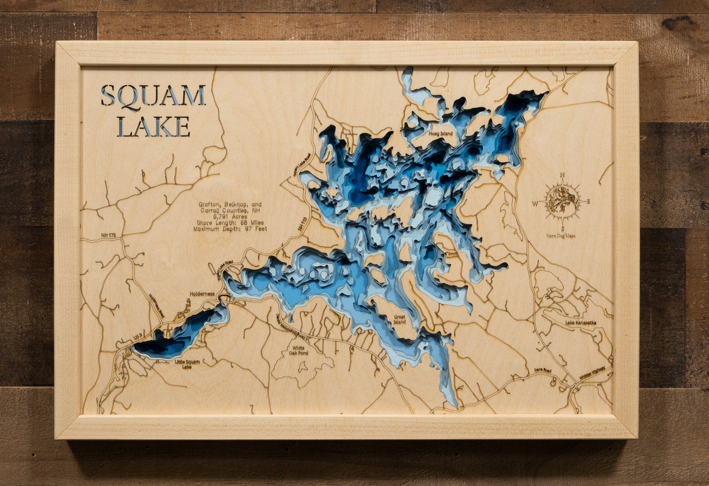 A laser-cut, layered wooden map of Squam Lake, located in Grafton, Belknap, and Carroll Counties, New Hampshire. The intricate shape of the lake, with its many islands contributing to its 68-mile shoreline, is clearly depicted within the central New Hampshire landscape. The varying depths of the lake, reaching a maximum of 97 feet, are represented by different layers of wood, creating a three-dimensional effect of this beautiful and well-known water body.
