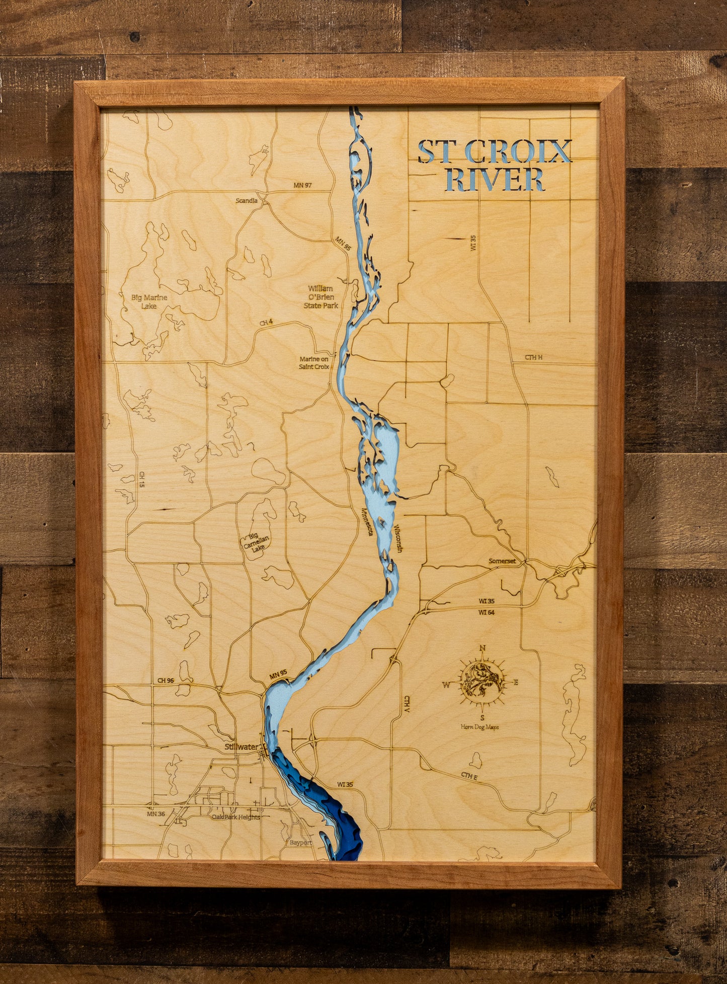 A framed wooden map of the St. Croix River, featuring a light natural wood background, engraved topography lines, a winding blue river, and a rustic brown wood frame, mounted on a dark wood plank wall.