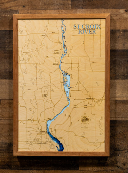 A framed wooden map of the St. Croix River, featuring a light natural wood background, engraved topography lines, a winding blue river, and a rustic brown wood frame, mounted on a dark wood plank wall.
