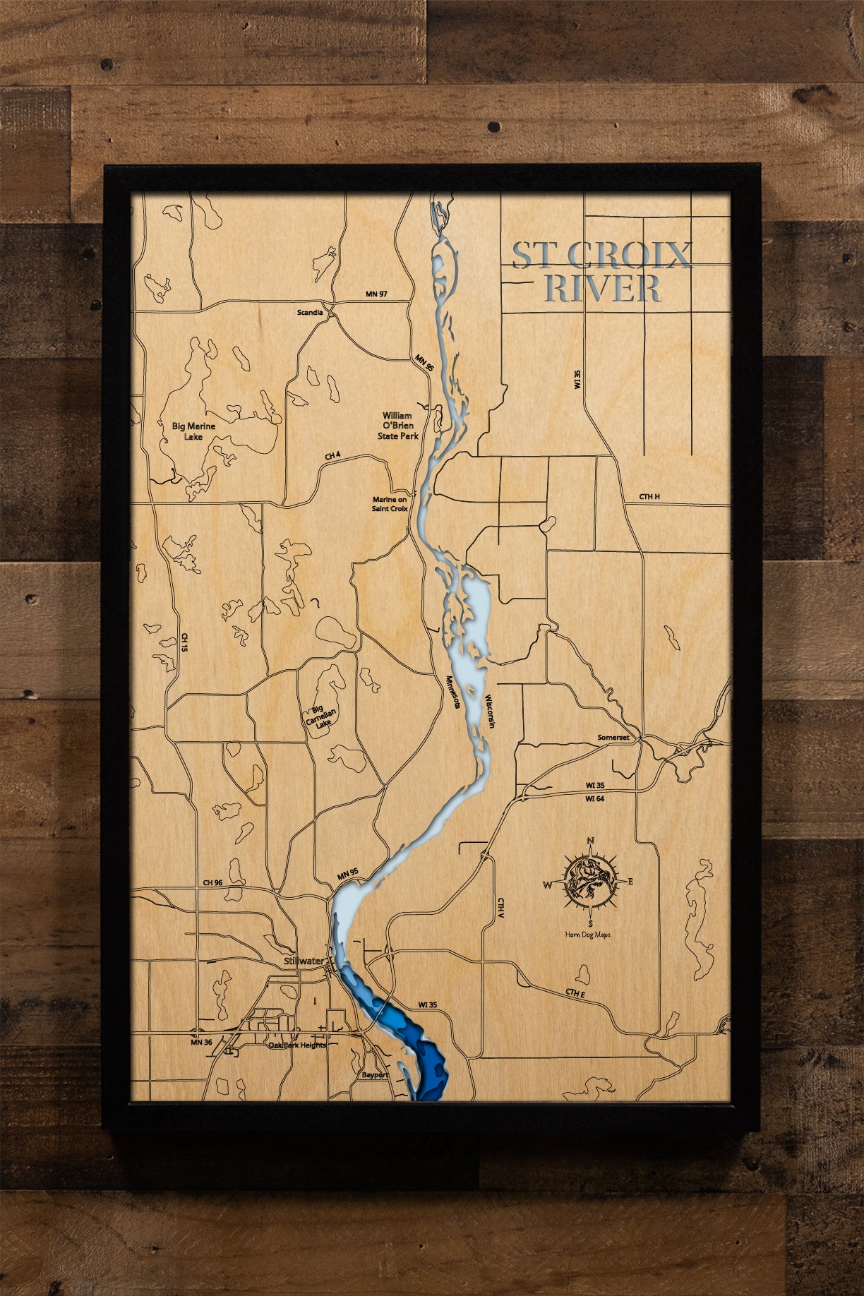 A framed wooden map of the St. Croix River, featuring a light natural wood background, engraved topographical details, and a blue river channel, mounted on a rustic dark wood plank wall.