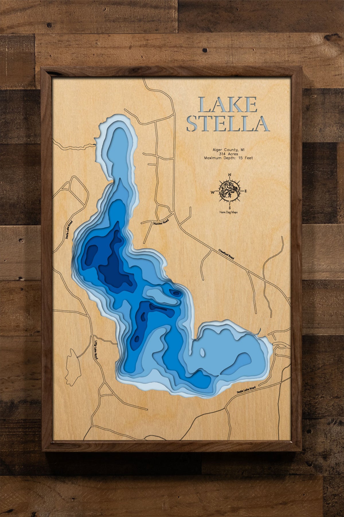 A precision laser-cut, multi-layered wooden map of Lake Stella in Alger County, Michigan, a 314-acre natural lake within the Hiawatha National Forest near Trenary featuring one island and one islet along its wooded shoreline. The natural wood top layer captures the lake's natural shoreline in fine detail, while a light-to-dark blue gradient conveys its shallow, peaceful character. A handcrafted statement piece for any Michigan UP cabin, National Forest retreat, or outdoor enthusiast's wall.