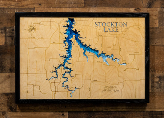 A framed wooden map of Stockton Lake, featuring a light natural wood background with etched contour lines, a prominent blue lake basin, and the words ' STOCKTON LAKE' in raised lettering. The map is housed in a thick black frame and mounted on a rustic dark wood plank wall.