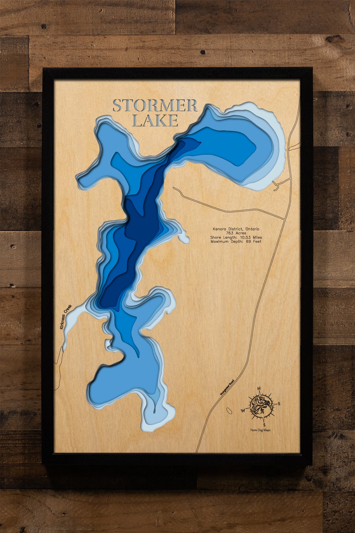 A highly detailed, laser-cut, and multi-layered wooden map of Stormer Lake, a natural lake in the Kenora District of Northwestern Ontario, Canada. The map shows the lake's distinct shape and its 10.53-mile shoreline, highlighting its connection to Kirkness Lake. The varying depths of the lake, which reaches a significant, unlisted maximum depth, are represented by different layers of wood.