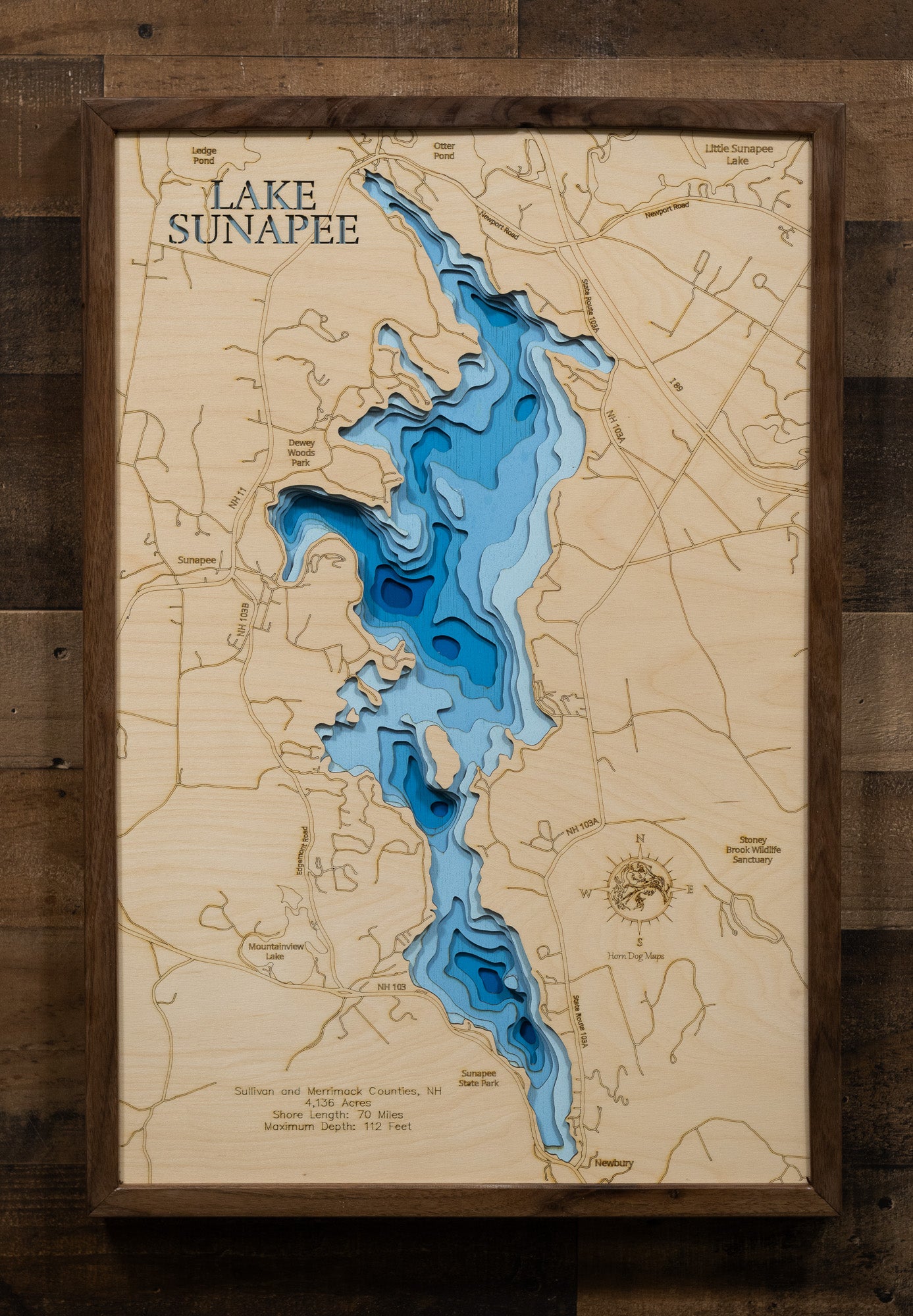 A laser-cut, layered wooden map of Lake Sunapee, located in Sullivan and Merrimack Counties, New Hampshire. The intricate shape of the lake, with its islands and varying shoreline, is clearly depicted within the western New Hampshire landscape. The varying depths of the lake, reaching a maximum of 112 feet, are represented by different layers of wood, creating a three-dimensional effect of this beautiful and popular water body.