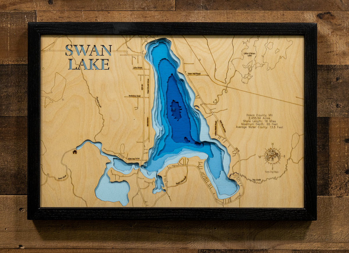 A framed 3D wooden depth map of Swan Lake, Washington, featuring layered wood pieces that create the lake's shape, with blue and light blue accents. The piece is mounted on a rustic dark wood wall.