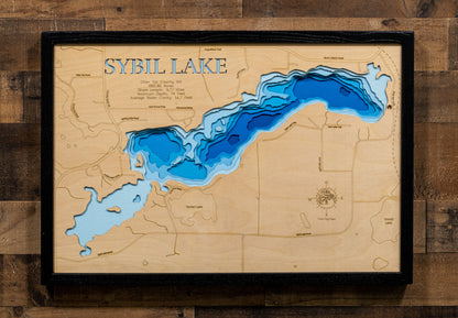 A framed wooden Sybil Lake depth map wall art piece, featuring layered blue and light blue water contours on a natural wood background, mounted on a rustic dark wood plank wall.