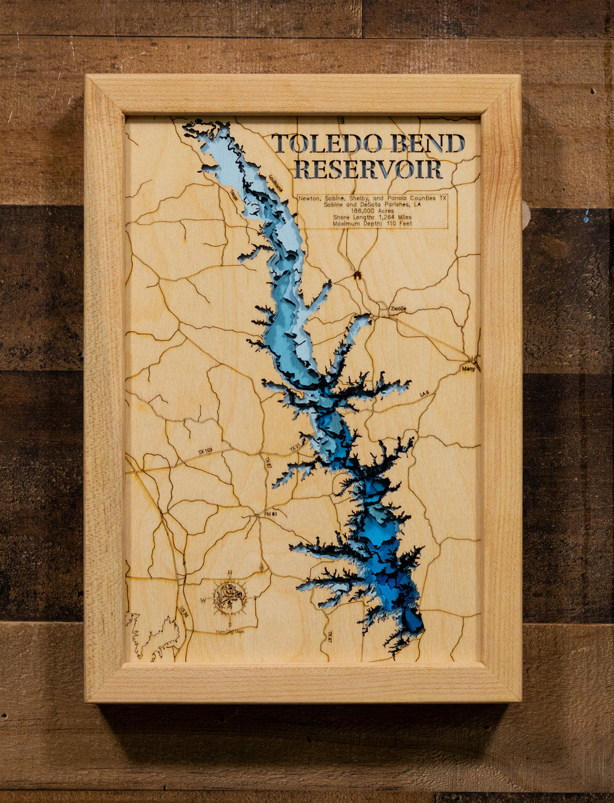 A laser-cut, layered wooden map of Toledo Bend Reservoir, located on the border of Louisiana and Texas. The expansive and intricate shape of this massive reservoir, stretching along the Sabine River, is clearly visible within the landscape of both states. The varying depths of the lake, reaching a maximum of 110 feet, are represented by different layers of wood, illustrating its underwater topography.