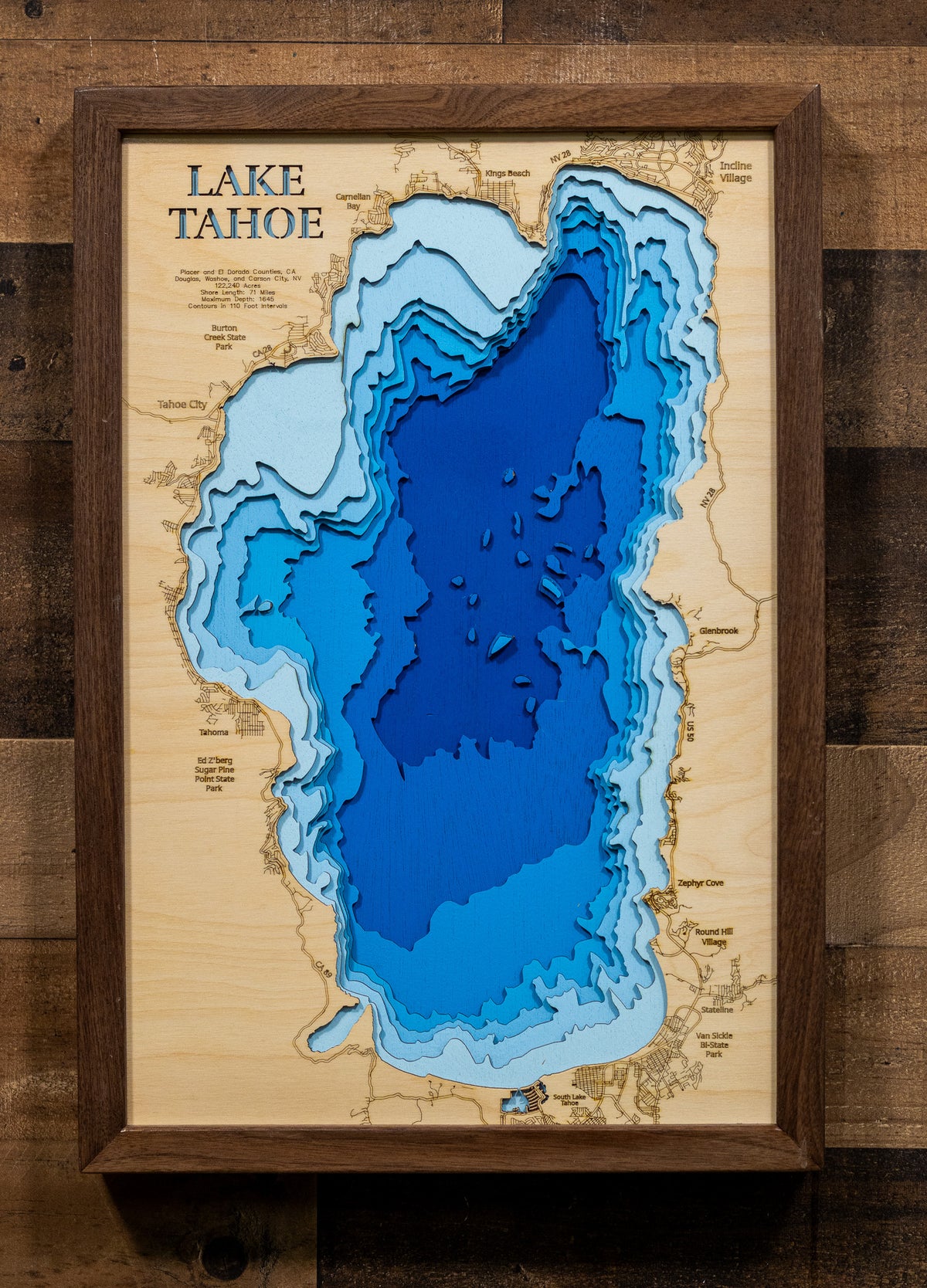 A framed 3D layered wood map of Lake Tahoe, showing depth contours in shades of blue on a natural wood background, mounted on a rustic dark wood wall.
