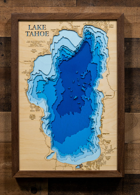 A framed 3D layered wood map of Lake Tahoe, showing depth contours in shades of blue on a natural wood background, mounted on a rustic dark wood wall.