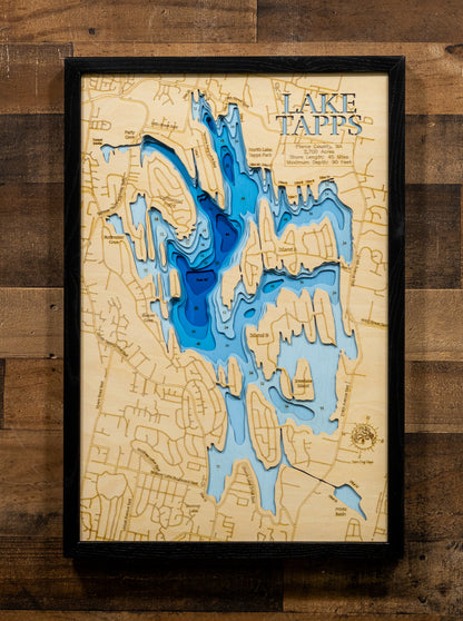 A framed 3D laser-cut wooden map of Lake Tapps, featuring layered blue water depths and a light beige wood background, mounted on a rustic dark wood plank wall.