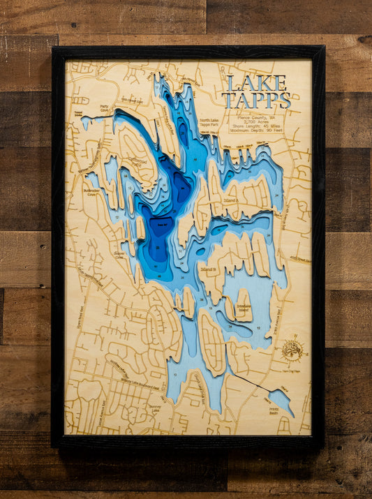 A framed 3D laser-cut wooden map of Lake Tapps, featuring layered blue water depths and a light beige wood background, mounted on a rustic dark wood plank wall.