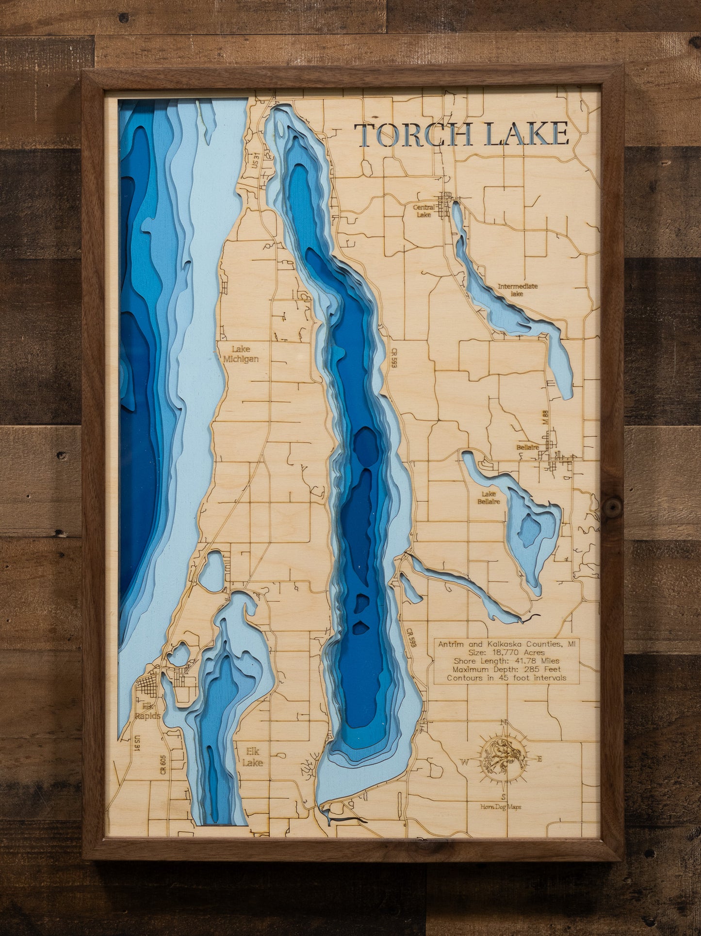 A laser-cut, layered wooden map of Torch Lake, located in Antrim and Kalkaska Counties, Michigan. The elongated and distinctive shape of the lake is clearly visible, highlighting its significant length within the Northern Michigan landscape. The varying depths of the lake, down to its impressive maximum, are represented by different layers of wood, with contour lines indicating depth changes at 45-foot intervals.