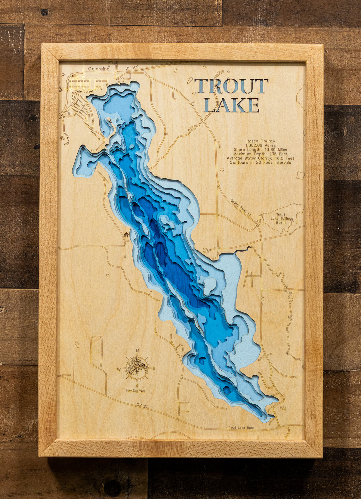 A framed wooden topographic map of Trout Lake, featuring layered blue and light blue wood sections representing the lake's depth, mounted on a rustic dark wood plank background.