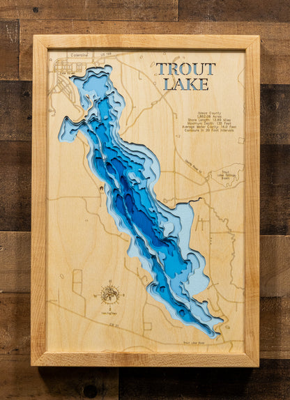 A framed wooden topographic map of Trout Lake, featuring layered blue and light blue wood sections representing the lake's depth, mounted on a rustic dark wood plank background.
