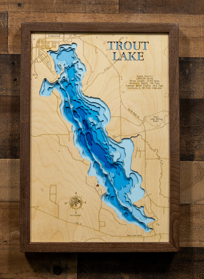 A framed wooden depth map of Trout Lake, featuring layered blue wood sections representing water depth on a light wood background, enclosed in a dark wood frame and displayed against a rustic wooden wall.