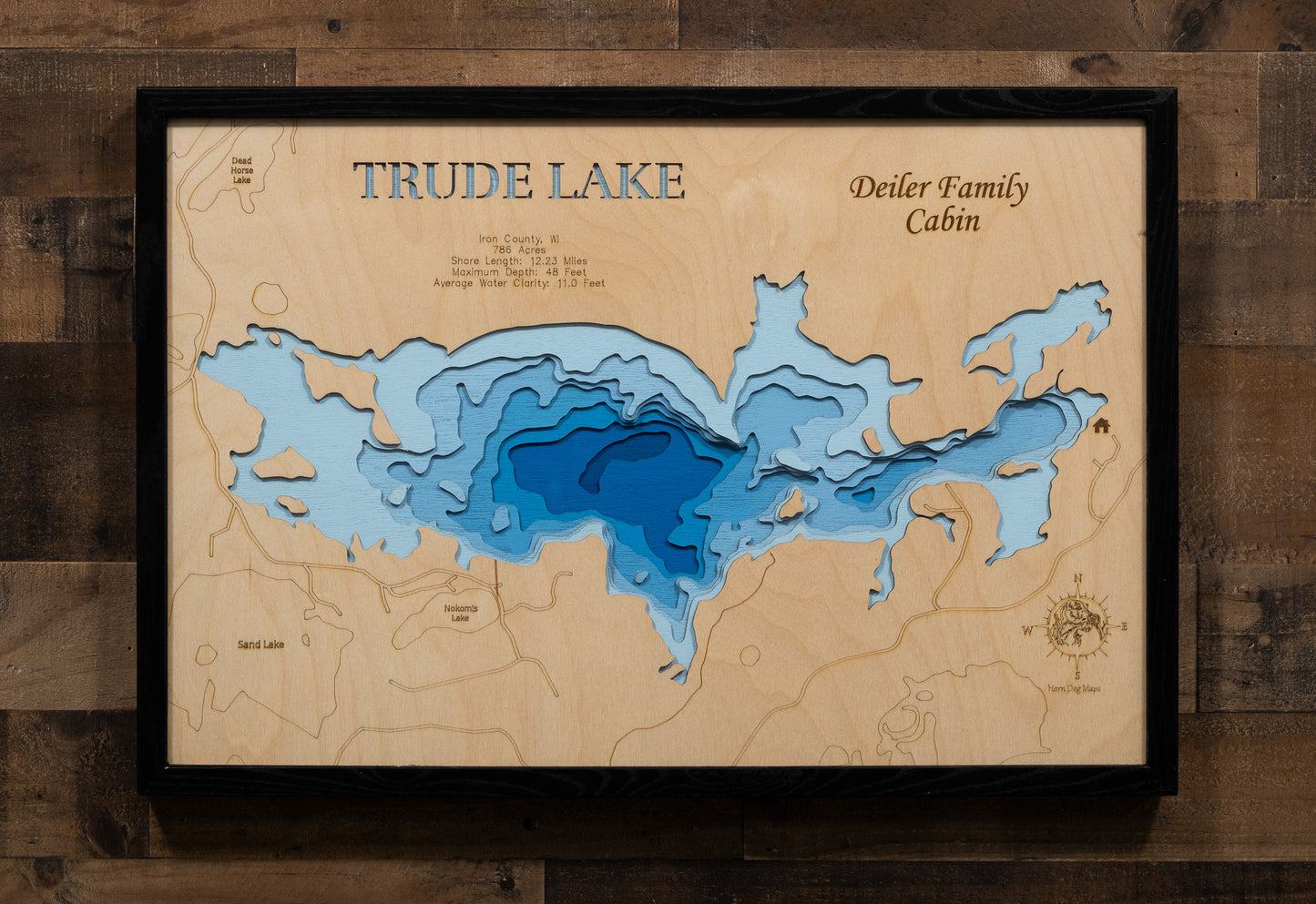 A highly detailed, laser-cut, and multi-layered wooden map of Trude Lake, a natural lake in Iron County, Wisconsin. The map shows the lake's distinct shape and its 12.23-mile shoreline. The varying depths of the lake, reaching a maximum of 48 feet, are represented by different layers of wood.