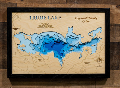 A framed 3D wooden topographic map of Trude Lake, featuring layered wood cutouts in shades of blue and white over a natural wood grain background, mounted on a rustic dark wood wall.