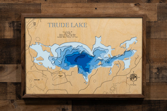 A highly detailed, laser-cut, and multi-layered wooden map of Trude Lake, a natural lake in Iron County, Wisconsin. The map shows the lake's distinct shape and its 12.23-mile shoreline. The varying depths of the lake, reaching a maximum of 48 feet, are represented by different layers of wood.