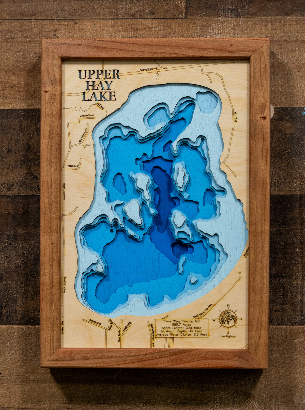A framed 3D wooden depth map of Upper Hay Lake, featuring layered blue wood sections representing water depth on a light wood background, enclosed in a medium brown wooden frame and mounted on a dark wood plank wall.