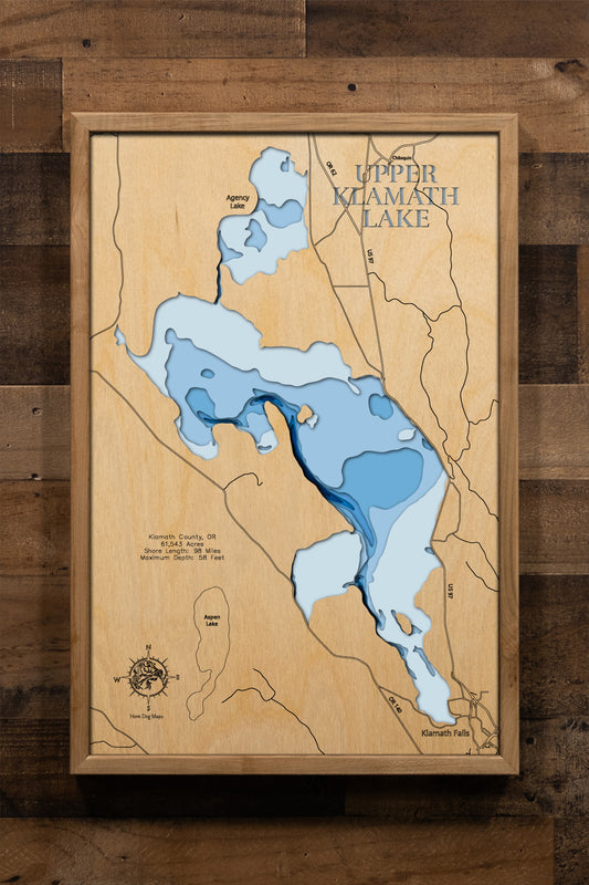 A meticulously crafted, multi-layered wooden topographic map of Upper Klamath Lake in Klamath County, Oregon. The laser-cut design showcases the lake's impressive 61,543-acre expanse stretching nearly 25 miles, the distinctive narrow western trench reaching 58-foot depths, the extensive marshlands and bays including Pelican Bay, and the 98 miles of intricate shoreline detail.
