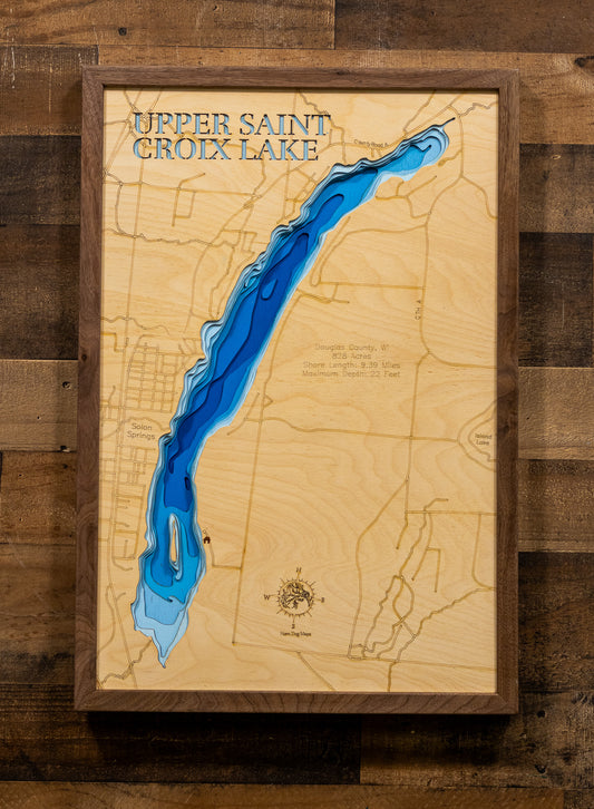 A framed wooden map of Upper Saint Croix Lake, featuring a natural light wood background, etched topographical details, and a raised blue lake channel. The piece is housed in a dark brown wood frame and mounted on a rustic dark wood plank wall.
